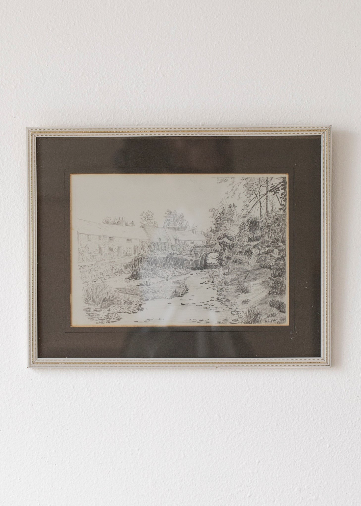 Hoppe Shoppe vintage pencil drawing of a stone bridge and thatched cottages in a decorative frame on a white wall.