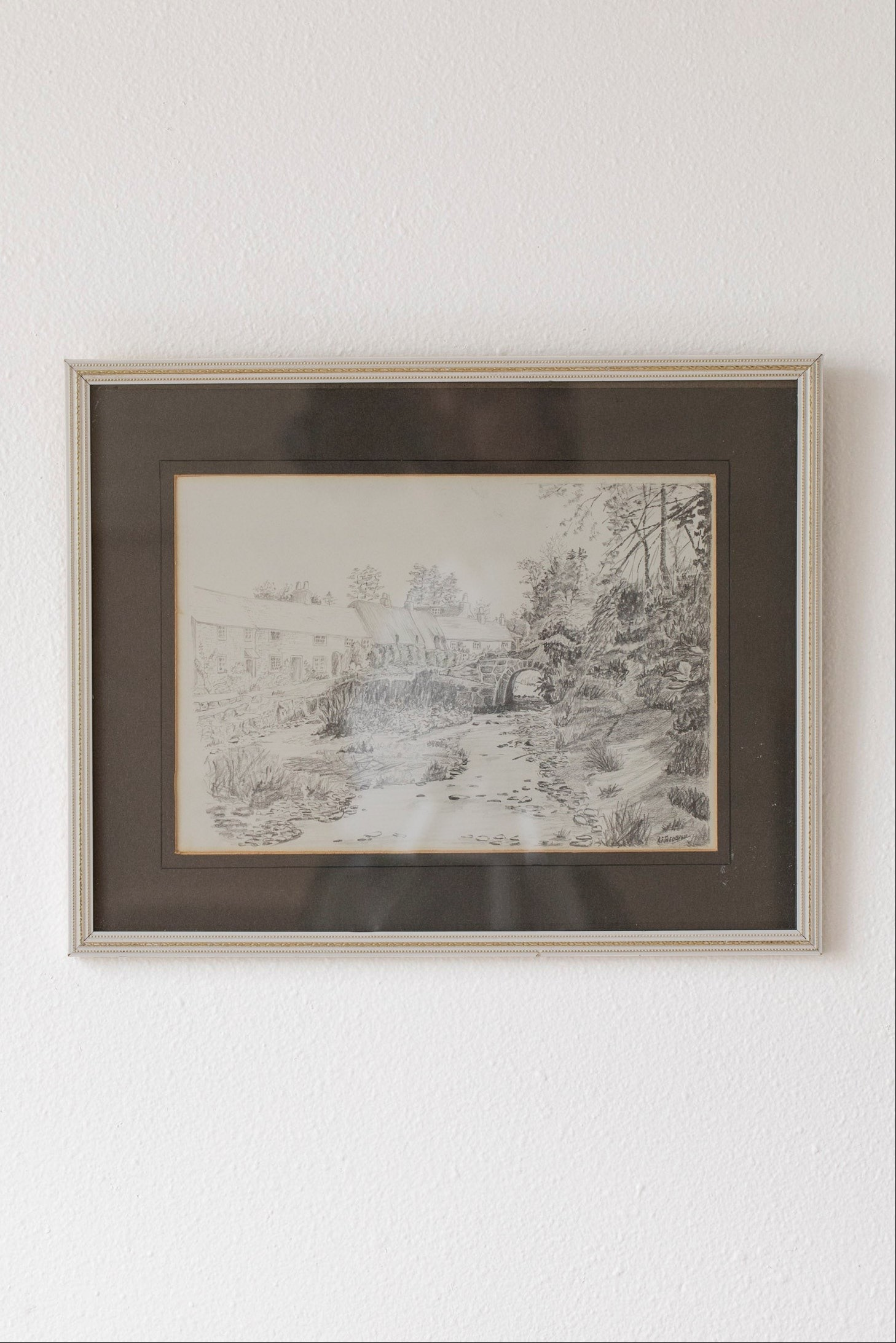 Hoppe Shoppe vintage pencil drawing of a stone bridge and thatched cottages in a decorative frame on a white wall.